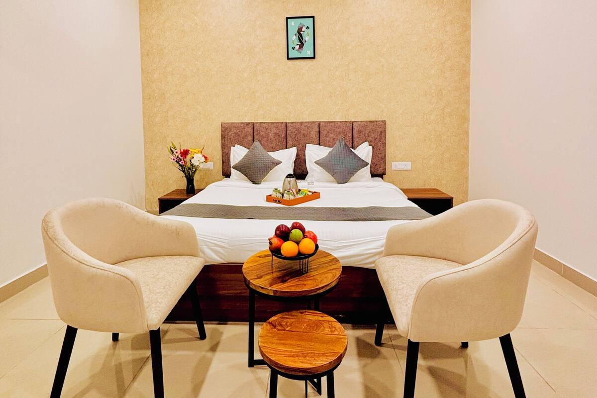 Quarto de hotel com uma cama, duas poltronas, uma pequena mesa de madeira com frutas e um quadro na parede.