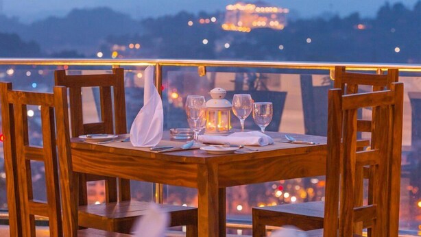 A wooden dining table set with glasses, a candle, and a napkin on a balcony with a view of a cityscape at dusk.