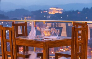 A wooden dining table set with glasses, a candle, and a napkin on a balcony with a view of a cityscape at dusk. - Sandriana Lake view (Kandy)