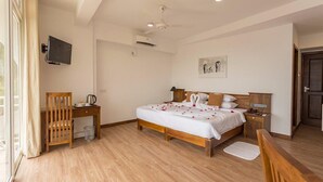 A hotel room with a wooden bed, a desk, a chair, a bedside table, a TV mounted on the wall, and a painting on the wall.