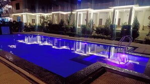 Una piscina con superficie de agua cristalina, rodeada por una zona junto a la piscina bien iluminada, con un camino empavedado y una escalera metálica.