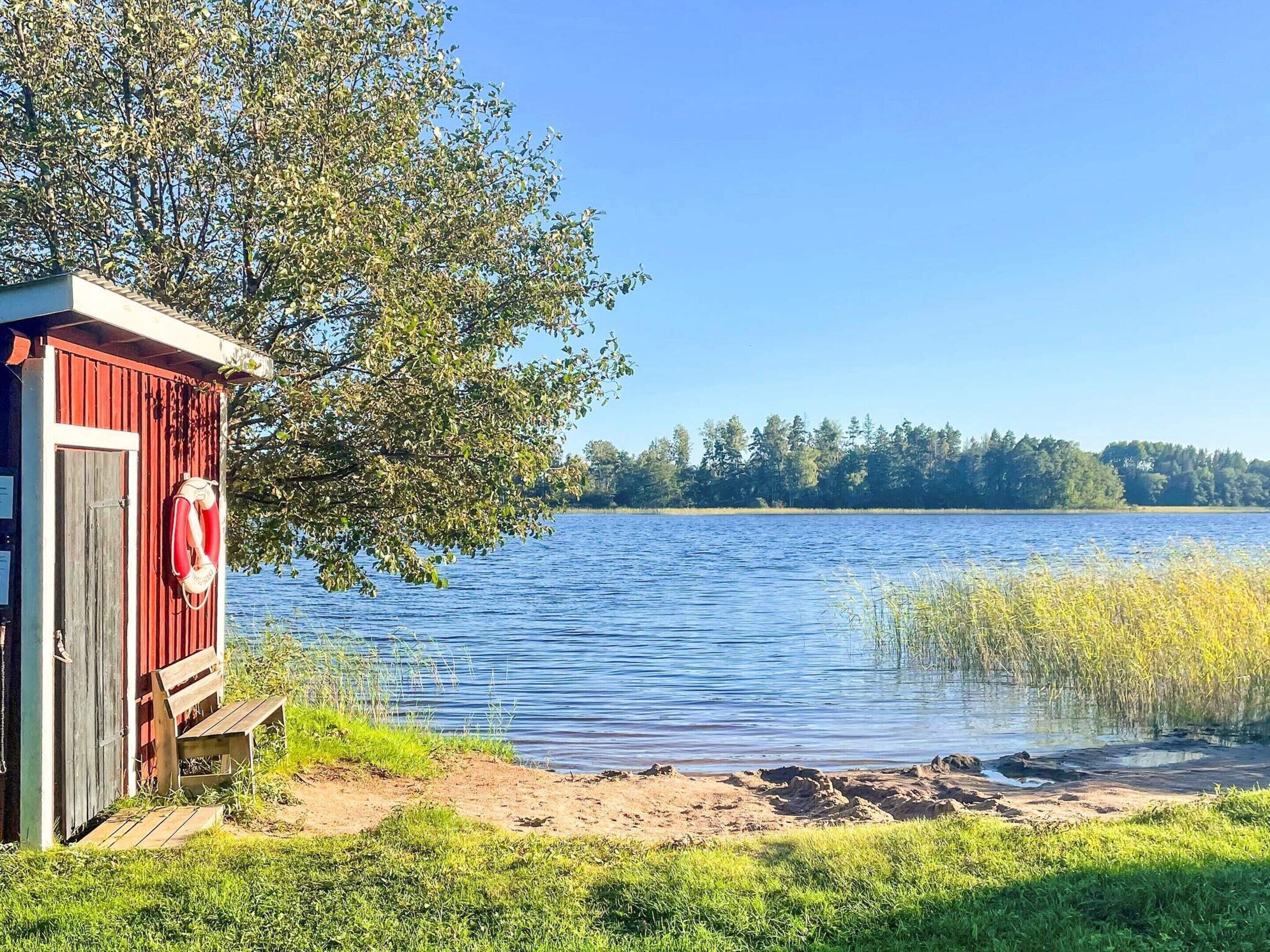 Un cobertizo rojo de madera con un banco y un flotador colgado en la puerta, situado junto a un lago tranquilo con una orilla boscosa al fondo.