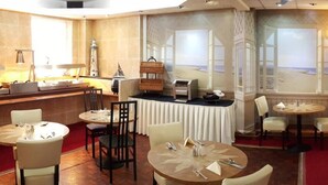 Ein Speisesaal eines Hotels mit runden Tischen, Stühlen, einer Buffetinsel und Blick auf den Strand durch große Fenster.
