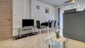 Una sala de estar moderna con un mueble de televisión de vidrio, comedor con sillas, un reloj y un refrigerador.