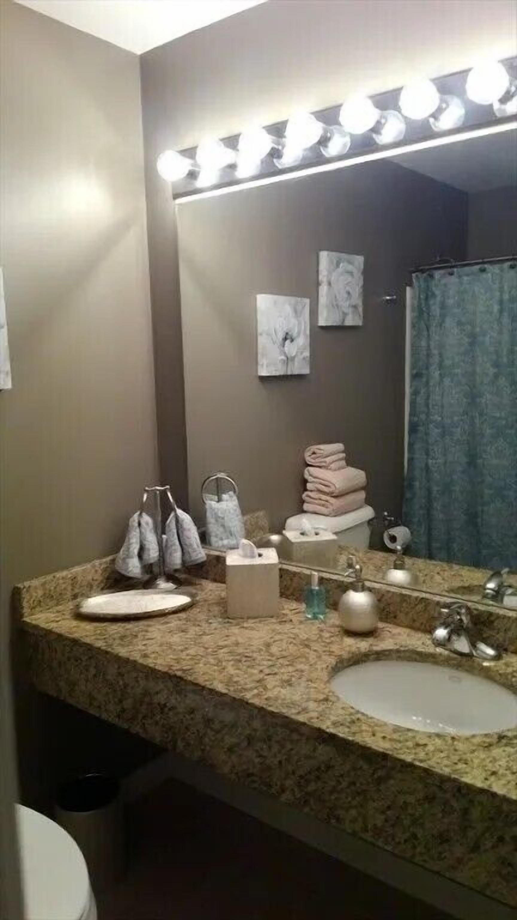 A bathroom with a granite countertop, two sinks, a mirror with lights, towels, and a shower curtain.