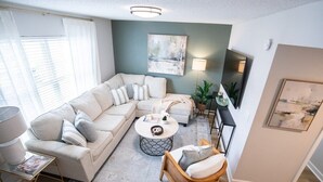 Un salon moderne avec un canapé d’angle, une table basse ronde et un fauteuil en osier.