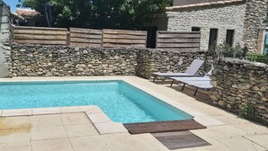 Une piscine rectangulaire aux murs en pierre et aux clôtures en bois, entourée d’une terrasse carrelée avec des chaises longues.