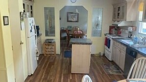 Uma cozinha compacta com piso de madeira, uma área de refeições com uma mesa pequena e cadeiras, e uma geladeira visível.