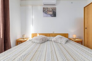 Sängyssä on kuviollinen vuodevaatesetti, kaksi pehmeää tyynyä, seinälle asennettu ilmastointilaite, puinen pääty ja pieni maalaus, jossa on polkupyörä.
