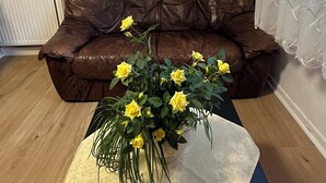 Un salón con un sofá de cuero marrón, una mesita con un jarrón de flores amarillas, un cuadro enmarcado en la pared y una ventana con cortinas de encaje.