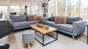Una sala de estar moderna con un sofá seccional gris, una mesa de centro de madera y un ventanal con vistas a árboles.