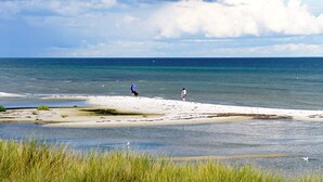 Una playa con dos personas caminando, dunas cubiertas de hierba y un cielo despejado.