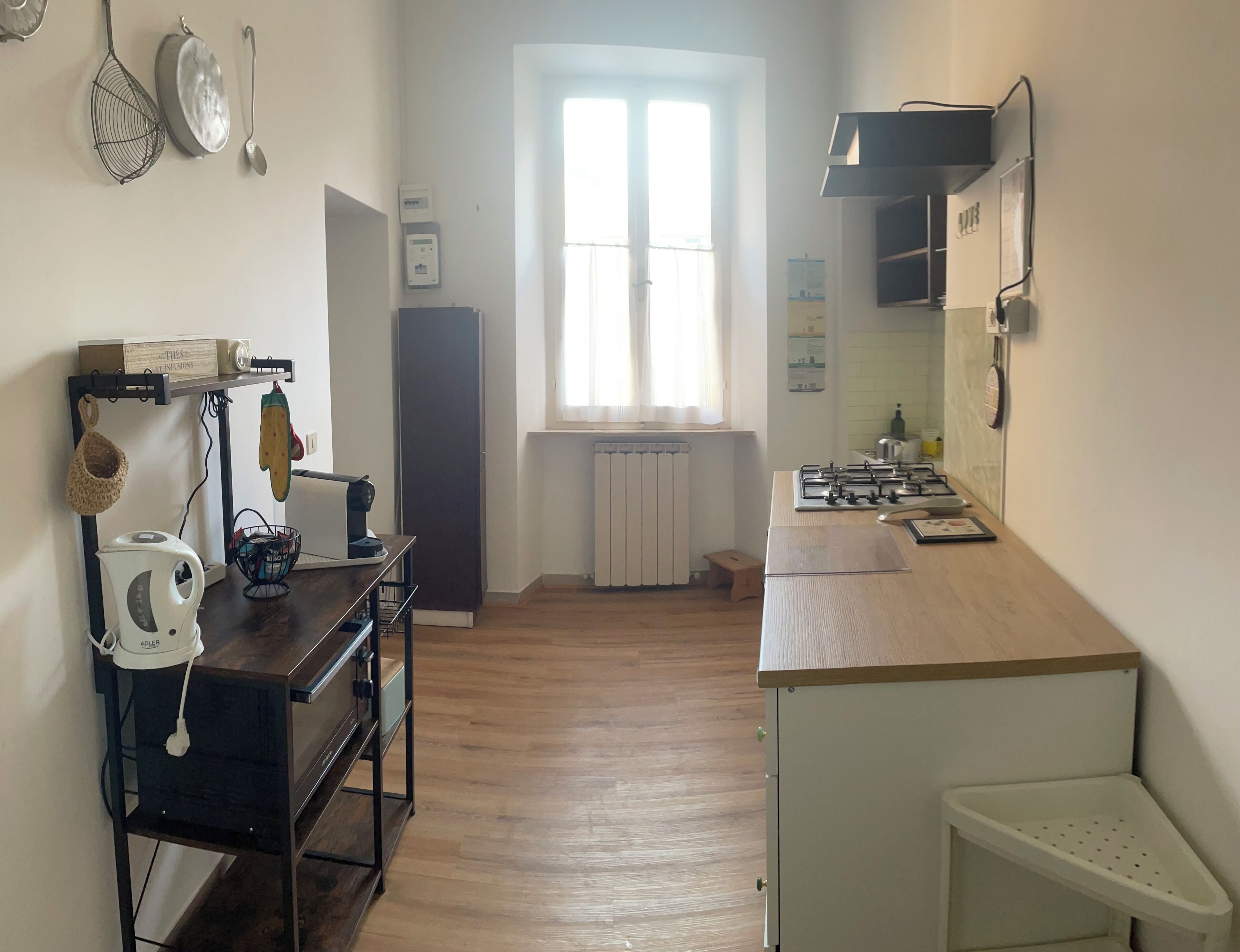 A kitchen with a stove, sink, and a window. There is a radiator under the window and a small stool.