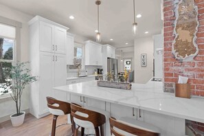 Une cuisine moderne avec des meubles blancs, un grand îlot central, des luminaires suspendus et un mur d’accent en briques.