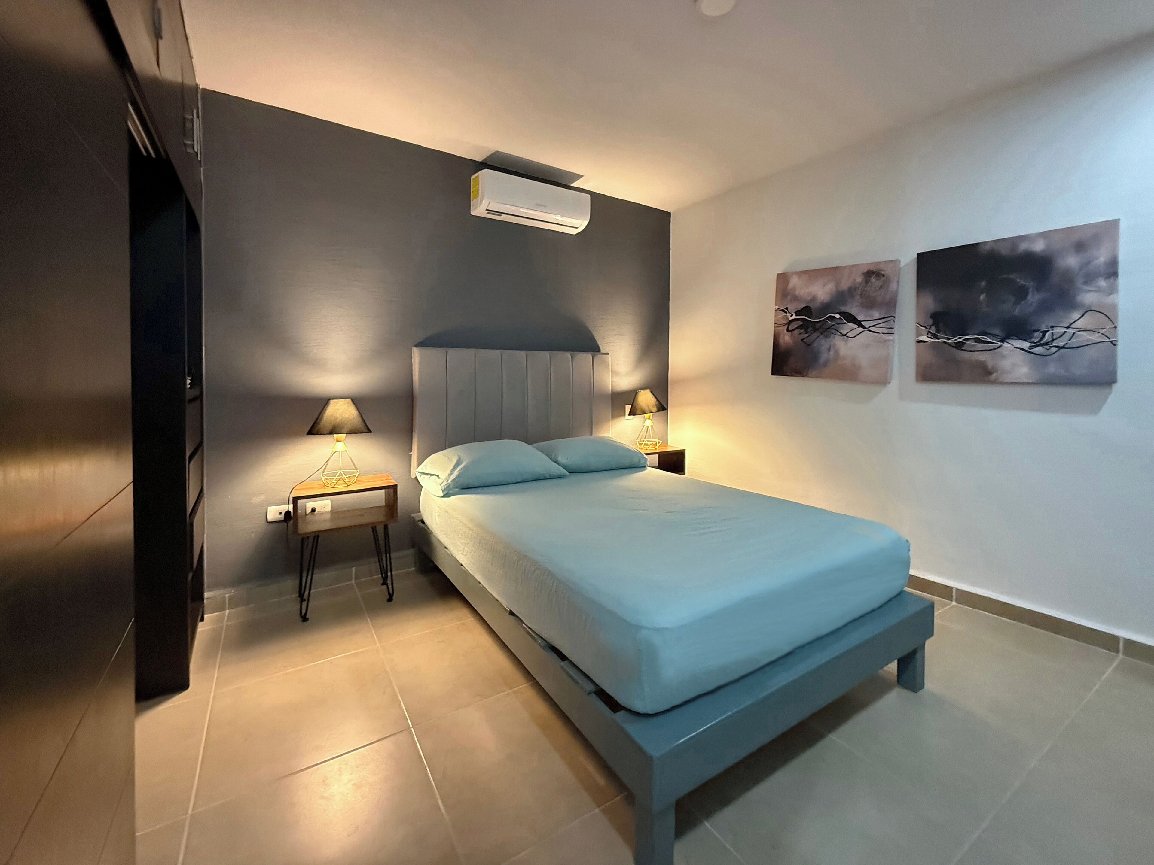 A modern bedroom with a bed, two bedside lamps, an air conditioner, and two wall art pieces.