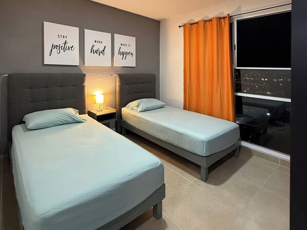 A hotel room with two beds, a nightstand with a lamp, and a window with orange curtains.