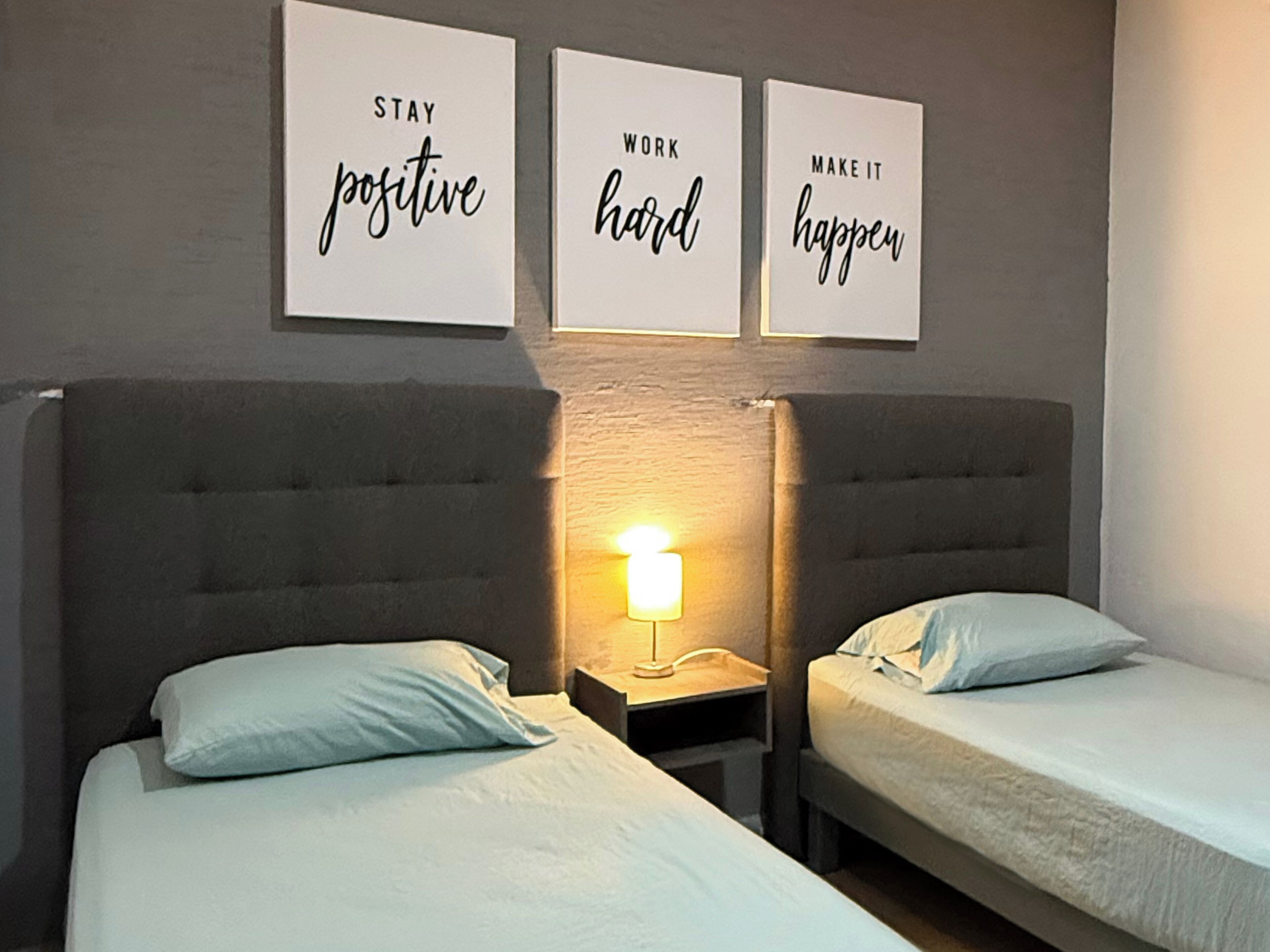 Une chambre d’hôtel avec deux lits, une table de chevet avec une lampe et trois affiches inspirantes accrochées au mur.