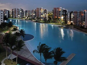 Une grande piscine à la forme incurvée, entourée de palmiers et de bâtiments d’appartements modernes.