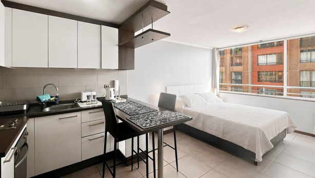 A compact hotel room with a kitchenette, a bed with white bedding, and a view of a brick building through a window.