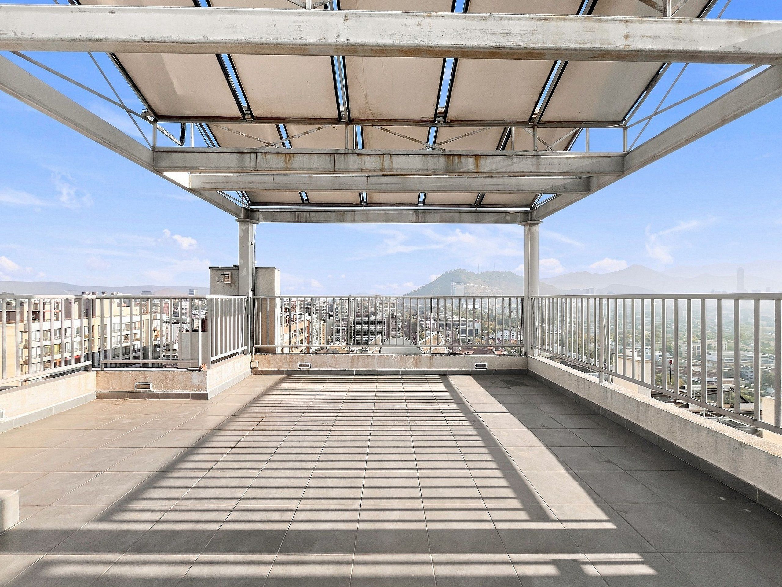 Una terraza abierta con vistas a un paisaje urbano, barandas de metal y piso de baldosas.