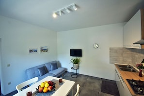 Une cuisine moderne avec un coin repas, un canapé gris, un téléviseur à écran plat et une horloge au mur.
