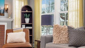 Un salón con un sillón de cuero marrón, un sofá gris, una mesa de centro de madera con revistas, una lámpara negra y una estantería con una planta en maceta.