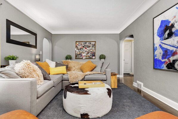 A modern living room with a grey sofa, a round ottoman, and a large abstract painting on the wall.