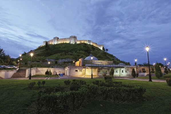 A historic castle on a hill with a modern building in the foreground, surrounded by well-maintained gardens and street lamps. - HOTEL GAZİ (Gaziantep)
