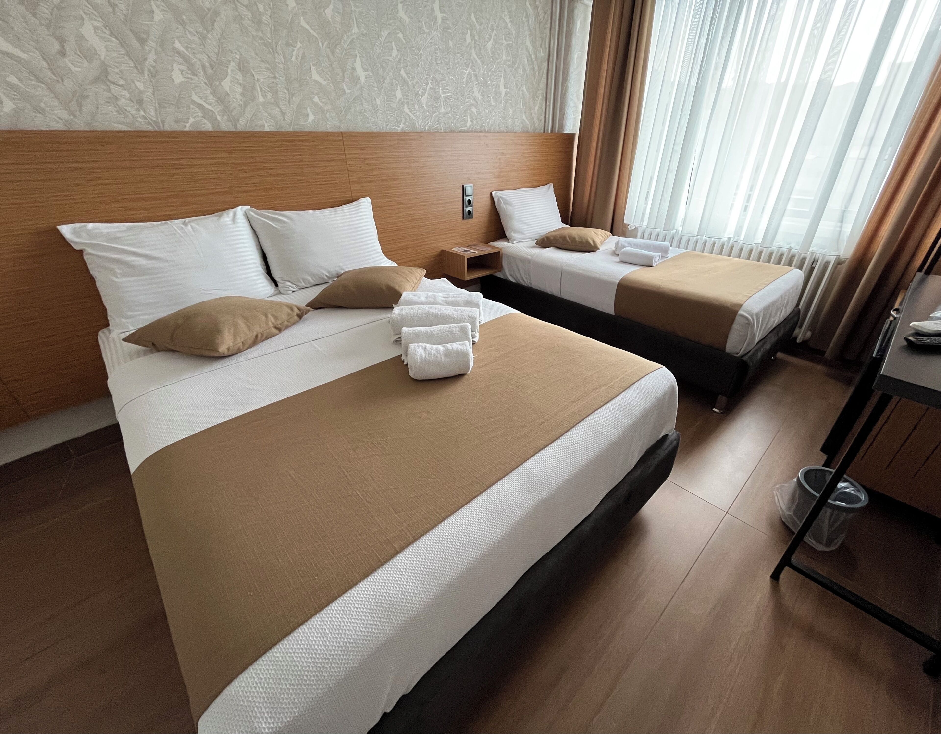 Een hotelkamer met twee bedden, elk voorzien van witte lakens en bruine sierkussens. Er is een houten hoofdbord, een klein houten nachtkastje en een raam met gordijnen.