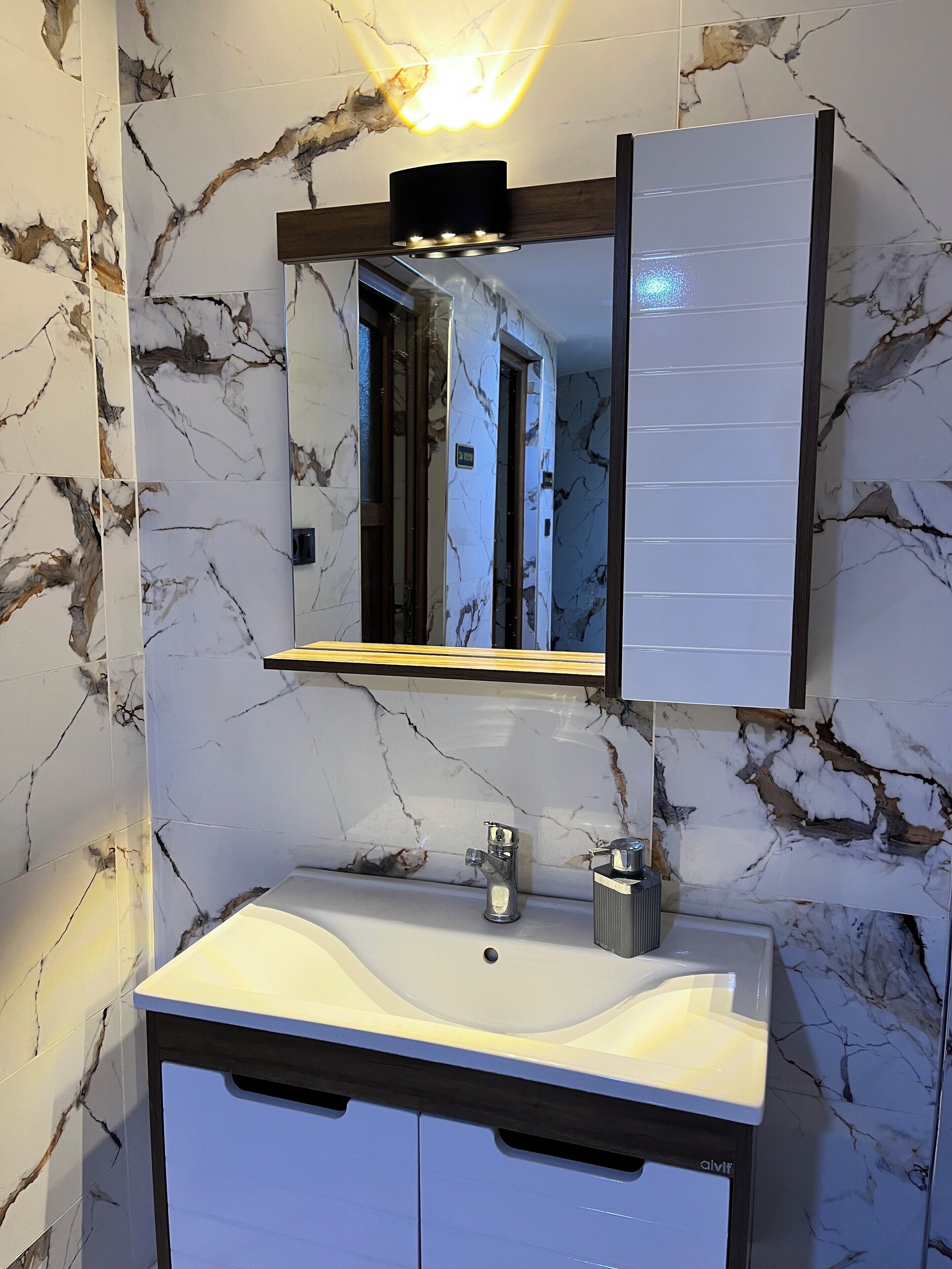 A bathroom with a marble backsplash, a white sink, a mirror with a black frame, and a shelf above the mirror.