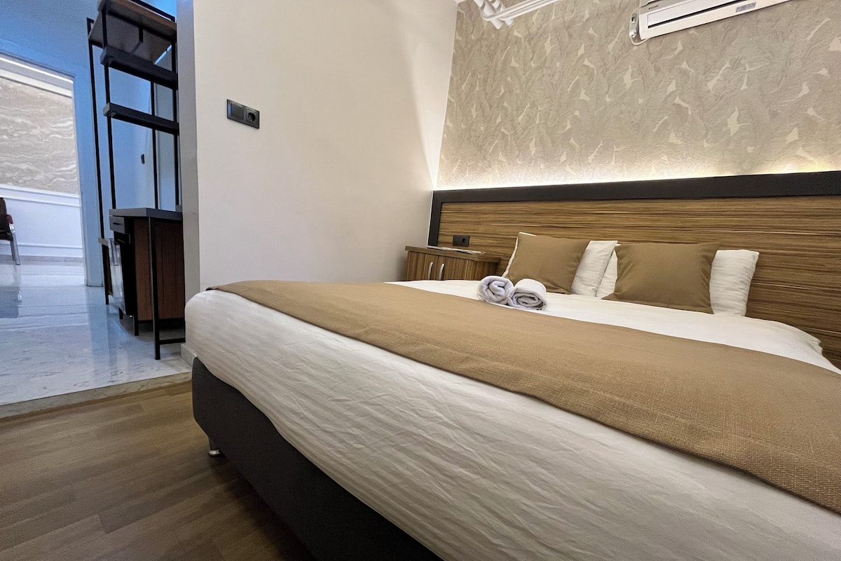A modern hotel room with a large bed, wooden headboard, and a view of the hallway.
