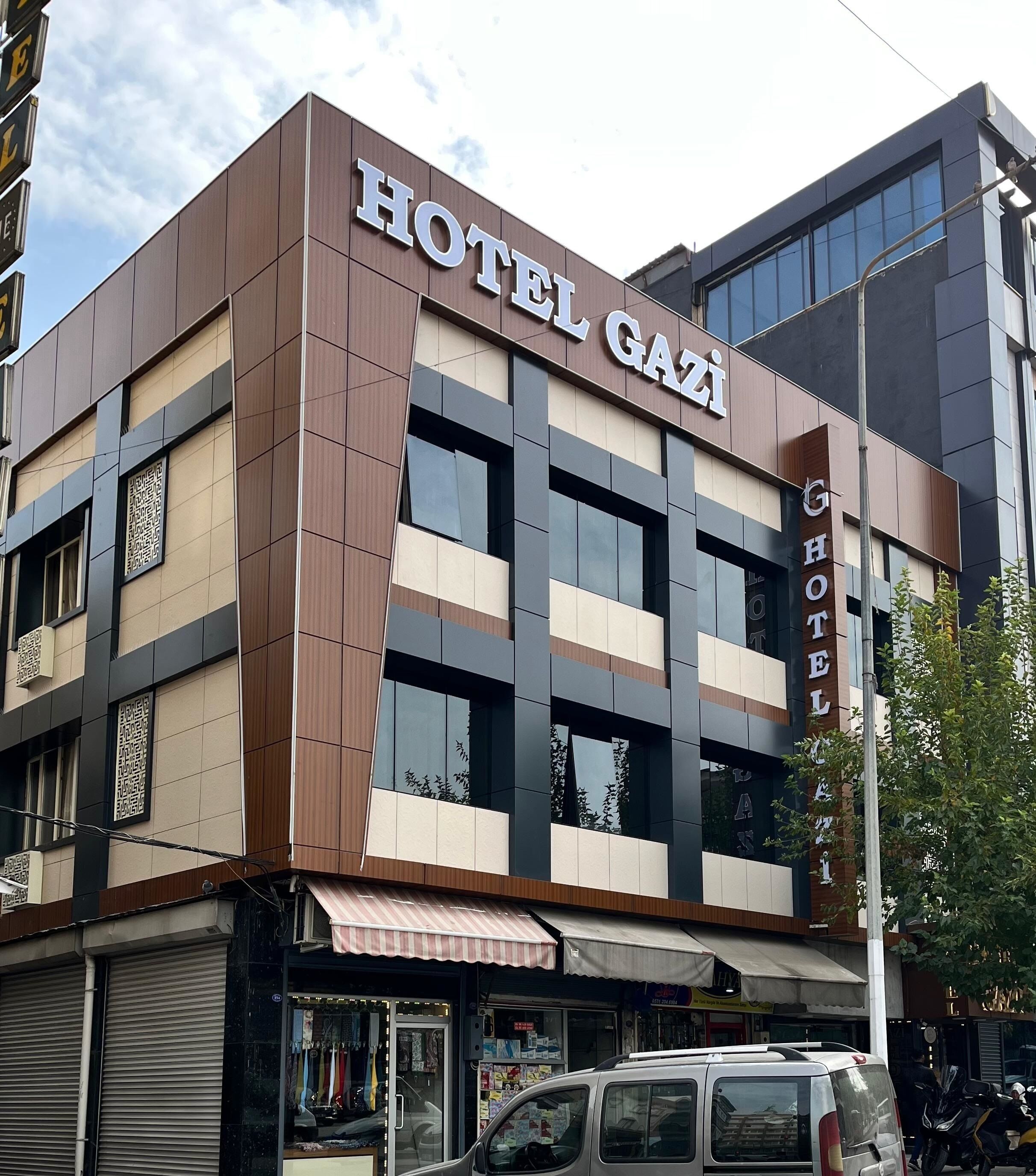 Un hotel llamado "HOTEL GAZI" con un diseño arquitectónico moderno que combina paneles marrón y beige, además de grandes ventanales.