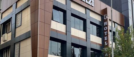 Un hotel llamado "HOTEL GAZI" con un diseño arquitectónico moderno que combina paneles marrón y beige, además de grandes ventanales.