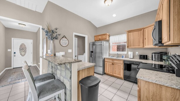 A modern kitchen with granite countertops, wooden cabinets, a stainless steel refrigerator, and a dishwasher. - Pet Friendly Home near Downtown Weatherford! Bring the Family, Relax & Enjoy! (Weatherford)