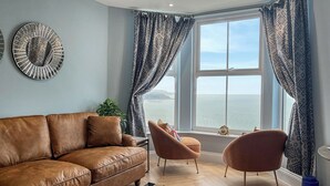 Ein Wohnzimmer mit braunem Ledersofa, zwei Sesseln, einem gemusterten Teppich und Meerblick durch große Fenster.