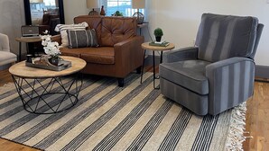 Una sala de estar con un sofá de cuero marrón, un sillón gris, una mesa de centro de madera y un tapete a rayas. Hay dos coronas colgadas en la pared y cuadros enmarcados con imágenes de vacas.