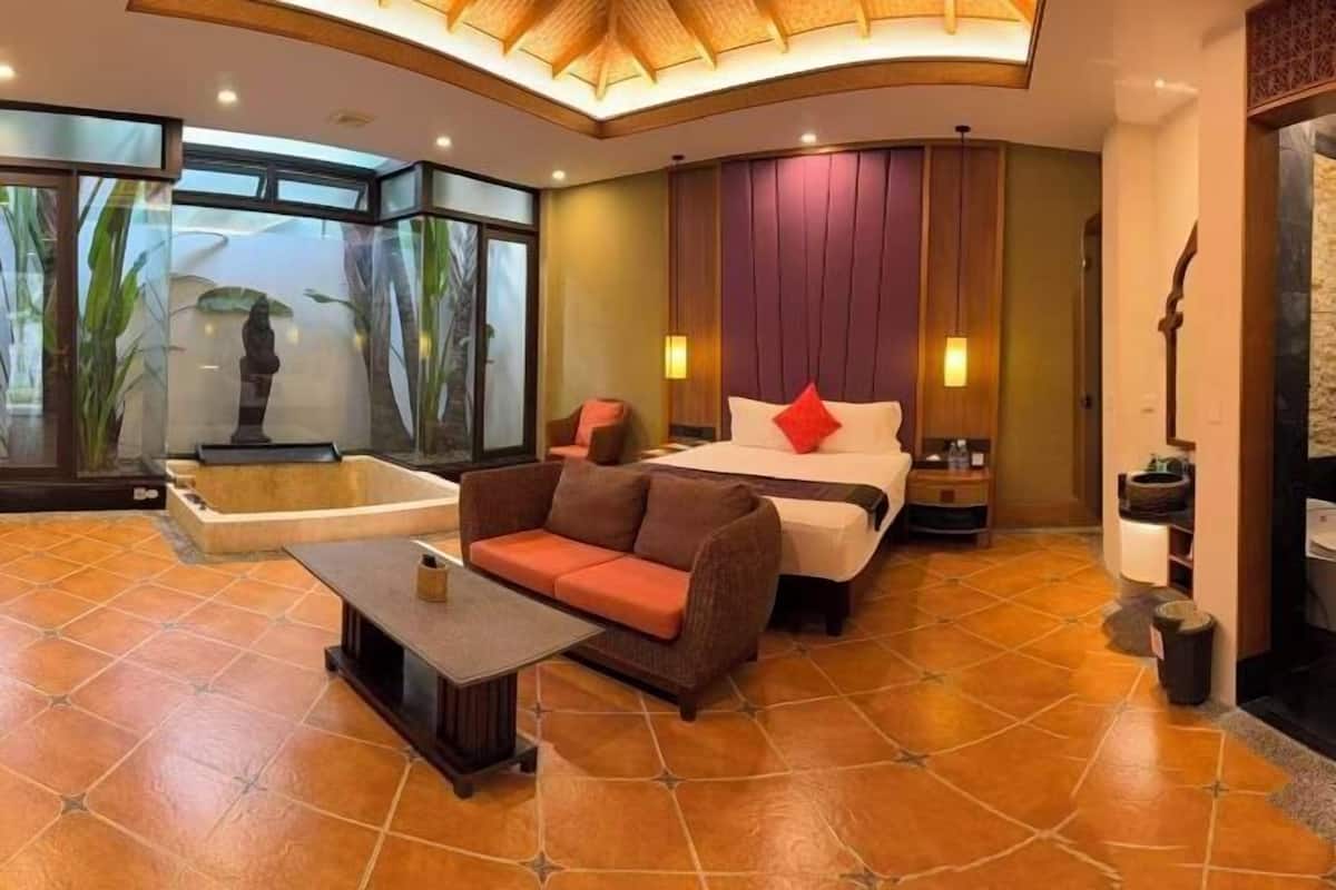 A modern hotel room with a large bed, a sofa, a small table, a bathtub, and a view of greenery through a glass wall.