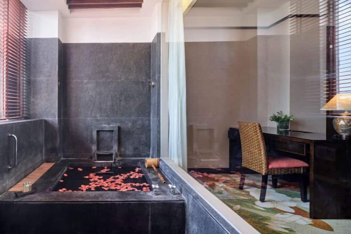 A bathroom with a bathtub, a shower, and a small step stool. There is a chair and a table with a lamp in the adjacent room.