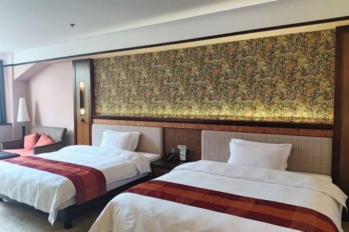 A hotel room with two beds, a large floral-patterned headboard, a nightstand, a lamp, and a chair.