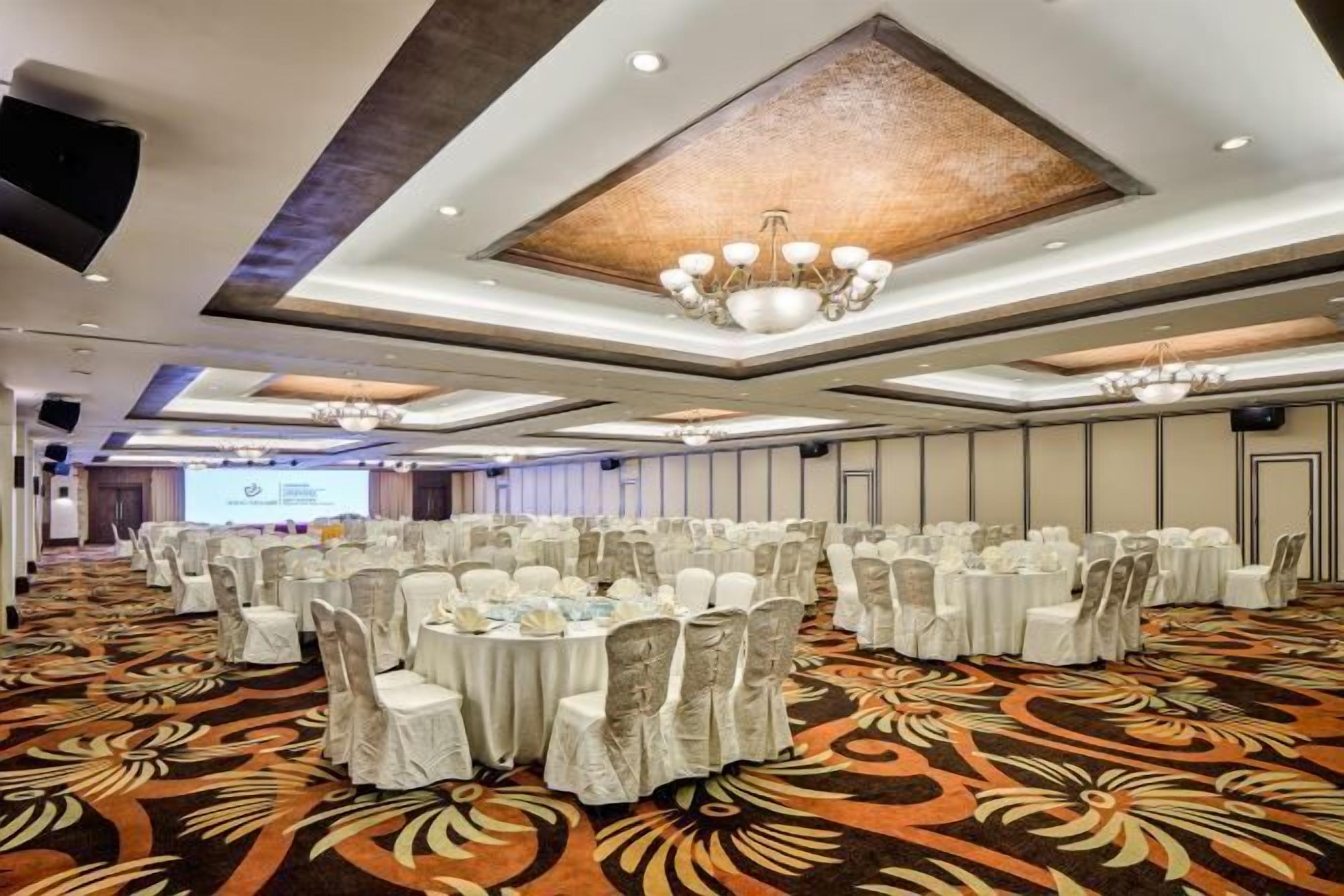 A large conference room with round tables, white tablecloths, and chairs covered in white fabric. The room has a patterned carpet and a modern ceiling design with recessed lighting and chandeliers.