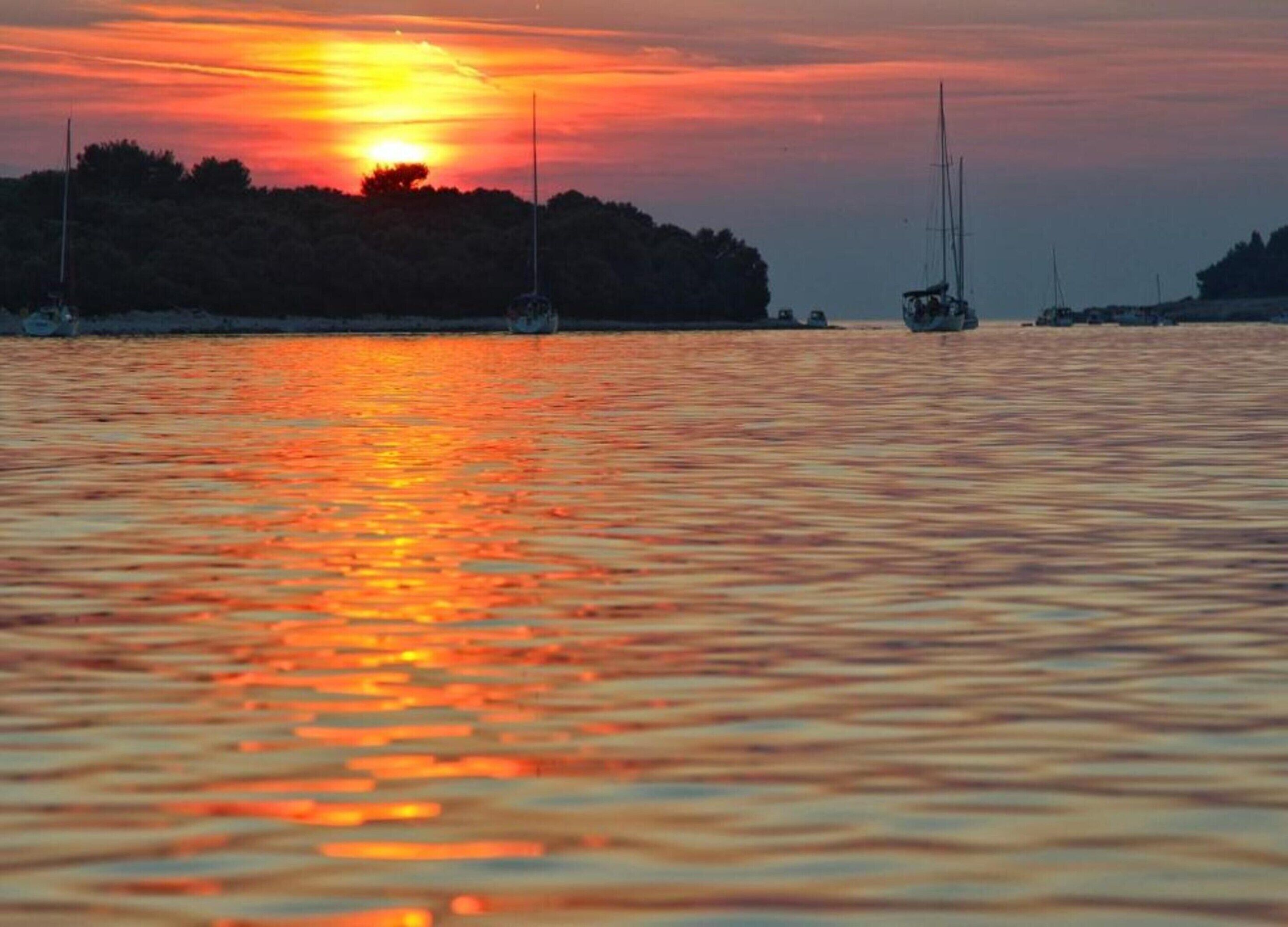 Een zonsondergang boven het water, met boten en een bebost landschap op de achtergrond.