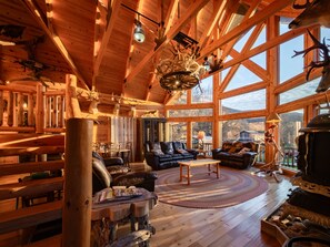 A log cabin interior with a large wooden ceiling, a fireplace, leather furniture, and a view of the outdoors.