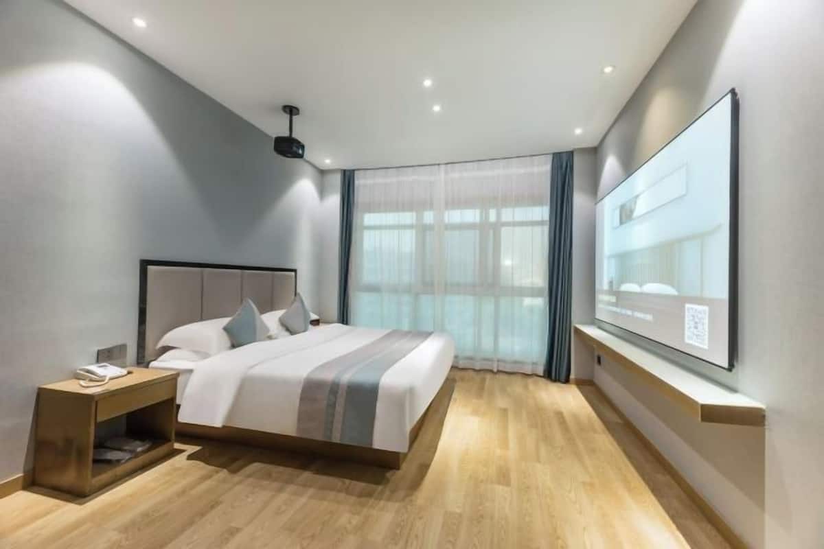 Una habitación de hotel moderna con una cama grande, una mesita de noche, un televisor de pantalla plana montado en la pared y un amplio ventanal con cortinas.
