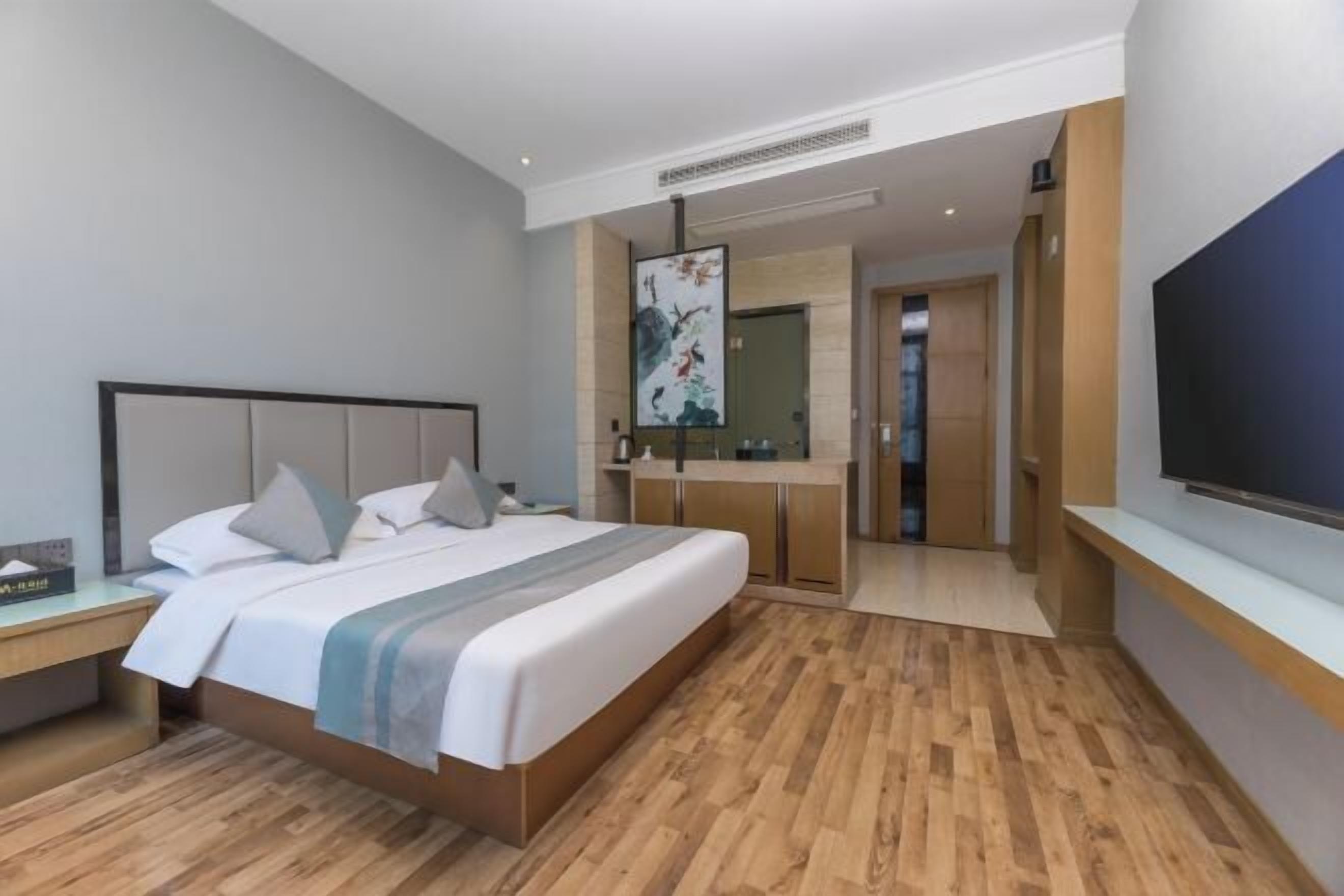 A modern hotel room with a large bed, wooden flooring, a TV, and a bathroom with a mirror and sink.