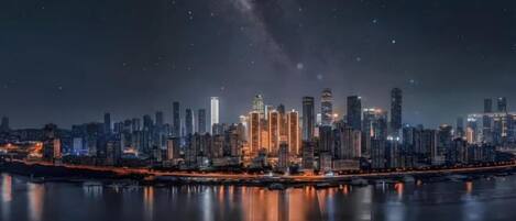 A nighttime cityscape with a prominent skyline of high-rise buildings, a calm body of water reflecting the lights, and a visible bridge connecting the two sides.