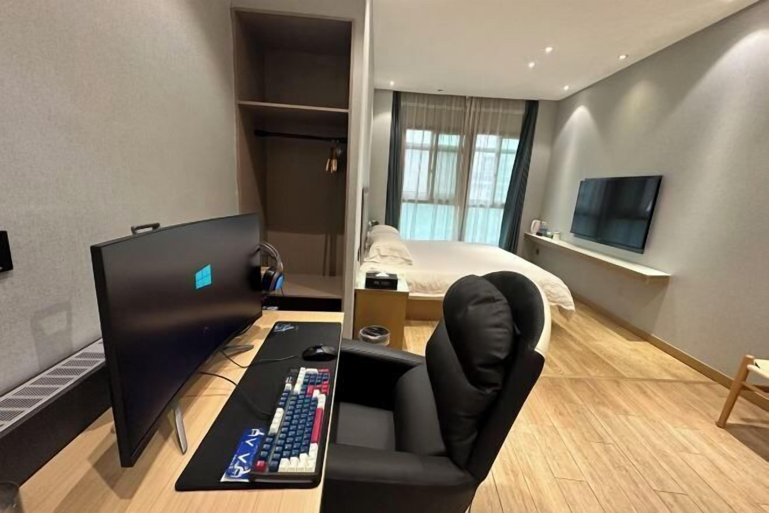 Une chambre d’hôtel moderne équipée d’un bureau, d’un ordinateur et d’un fauteuil de jeu. On y trouve un lit avec un téléviseur fixé au mur, ainsi qu’une fenêtre avec des rideaux.