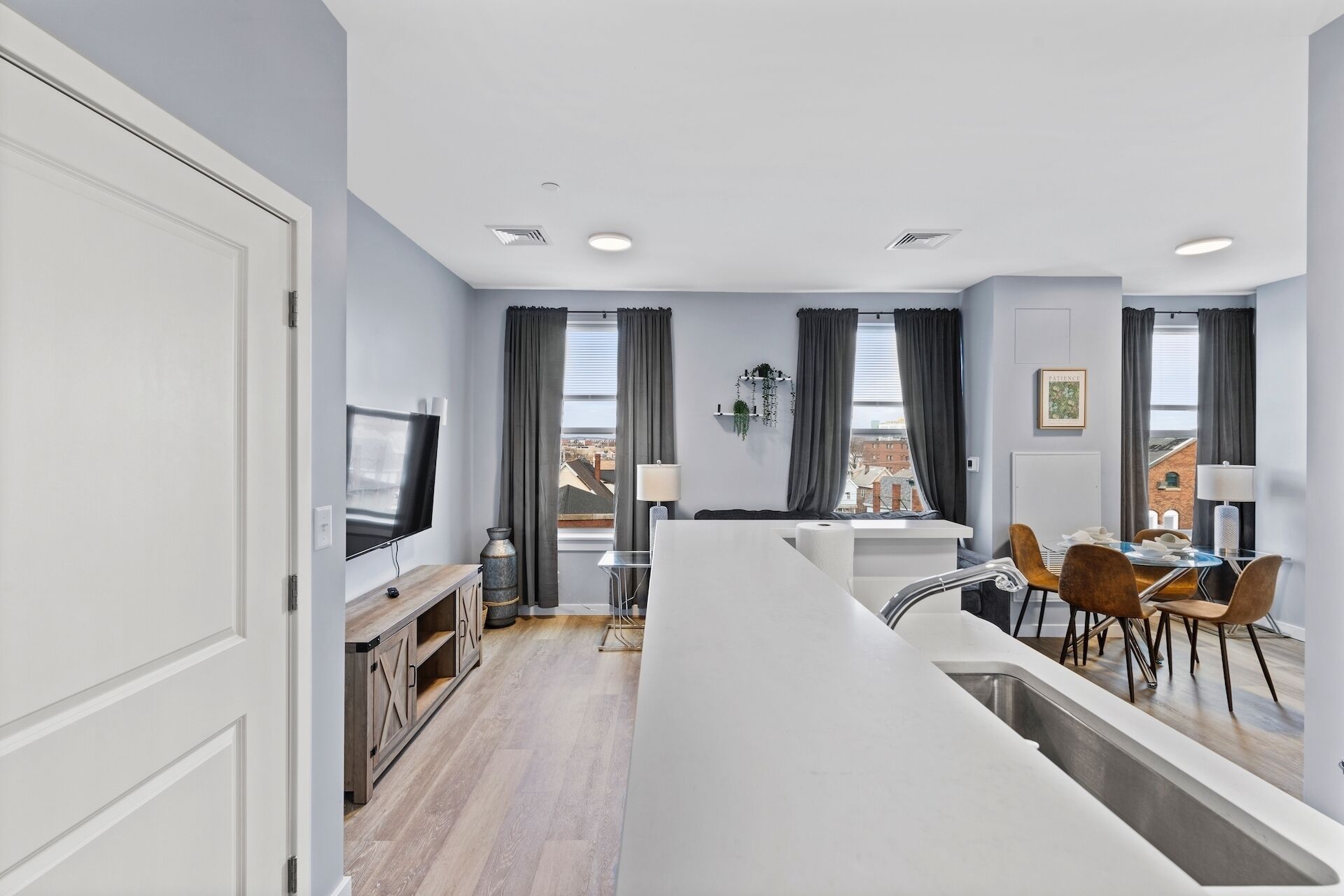 A modern kitchen with a central island, a dining area with chairs, a TV mounted on the wall, and a window with curtains.
