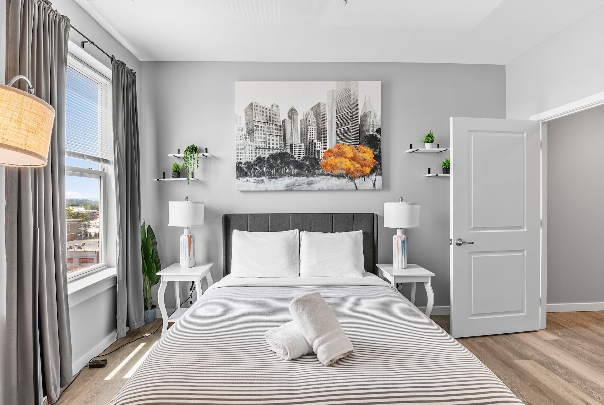 Un dormitorio moderno con una cama, lámparas de noche, un cuadro de un paisaje urbano y vistas de la ciudad a través de la ventana.