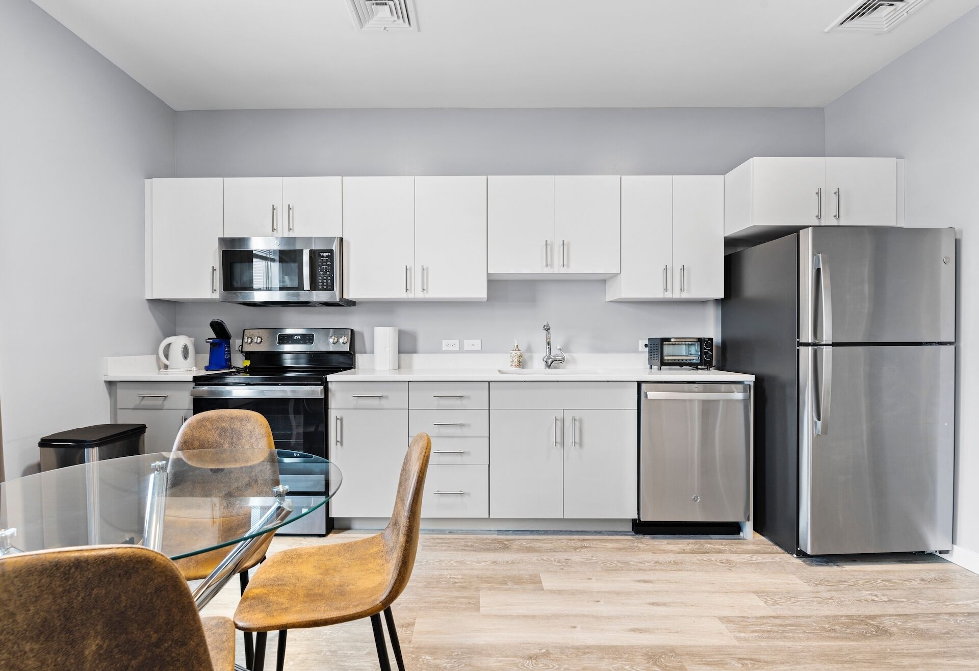 A modern kitchen with white cabinets, stainless steel appliances, a glass dining table, and wooden chairs.