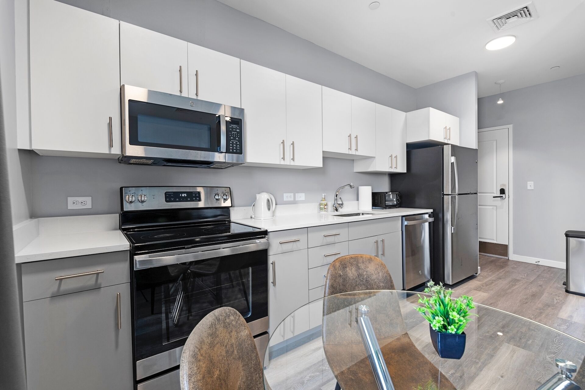 A modern kitchen with white cabinets, stainless steel appliances, a glass dining table, and a potted plant.